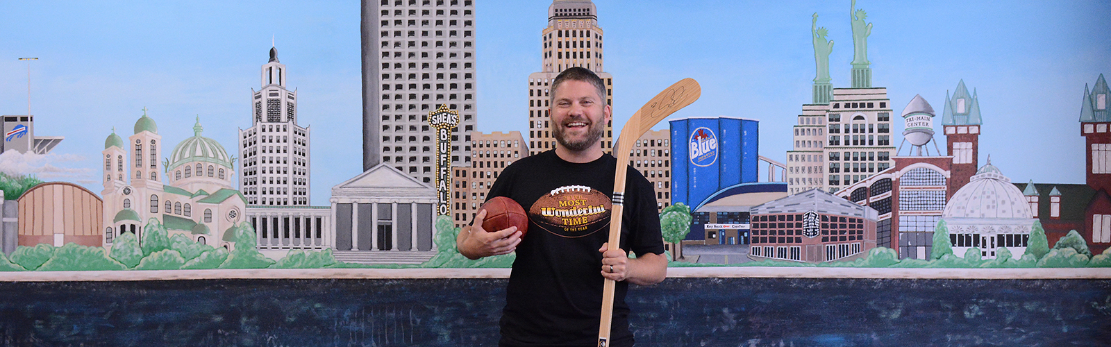 Sporting one of his T-shirt designs, 26 Shirts co-founder Del Reid stands in front of a mural of the Buffalo skyline at his office in the Tri-Main Center. Reid specializes in Buffalo-themed shirts.